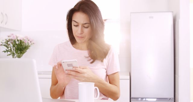 This image depicts a woman in casual clothing sitting in a bright, white kitchen. She is using a smartphone with a coffee mug nearby, suggesting she is multitasking and possibly working from home. This can be used in articles or advertisements related to remote work, home office setups, technology use in daily life, and balancing personal routines with professional tasks.