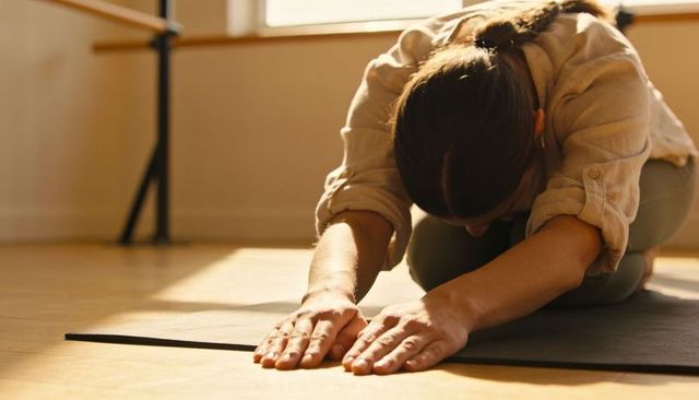 Kneeling woman stretching forward on yoga mat in sunlit studio with barre and hardwood