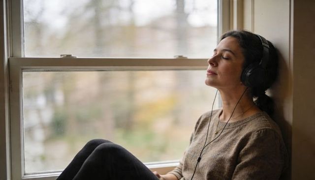 Woman listening on window seat wearing over-ear headphones, relaxing with eyes closed
