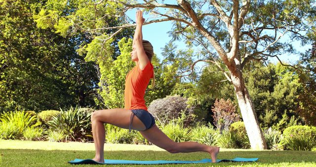 Young woman practicing yoga outside in a lush, green park. She is performing a lunge pose on a yoga mat, surrounded by trees and nature. Perfect for lifestyle, health and wellness, outdoor activities promotions, and fitness content.