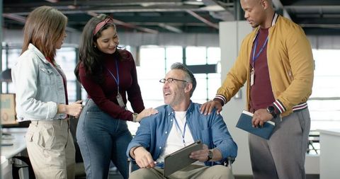 Diverse office team supporting colleague in wheelchair, collaborating on tablet in bright office