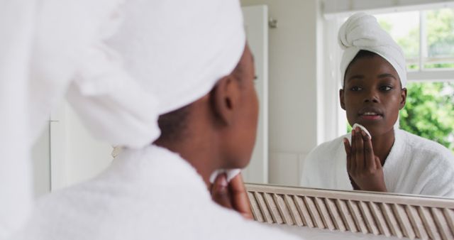 African american woman in bathrobe cleaning her skin with a cotton pad while looking in the mirror at home. lifestyle living self isolating in quarantine lockdown