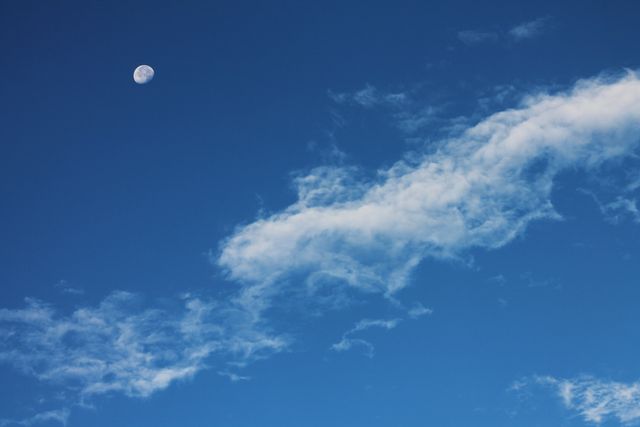 Beautiful daytime sky with a visible crescent moon surrounded by wispy white clouds against a blue background. Perfect for nature-themed blogs, peaceful scene backgrounds, meditation or relaxation content, and celestial illustrations.