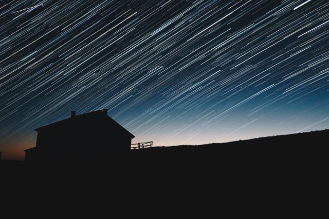 Star trails appear over a silhouette of a rural house, creating a striking composition. Ideal for use in materials about astronomy, rural life, night photography, or celestial studies. Captures the beauty and tranquility of countryside nights, perfect for nature and science content.
