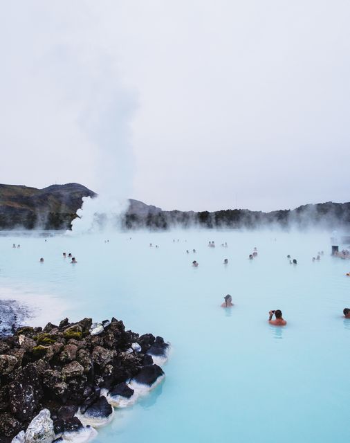 People are enjoying the Blue Lagoon's geothermal hot springs surrounded by volcanic rocks. Steam rises from the heated waters, creating a magical and relaxing atmosphere. This image can be used for promotional materials focused on travel, wellness tourism, Icelandic attractions, and nature retreats.