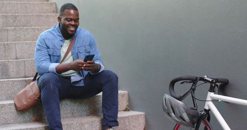 African American man sitting on steps checking smartphone beside parked bicycle and helmet