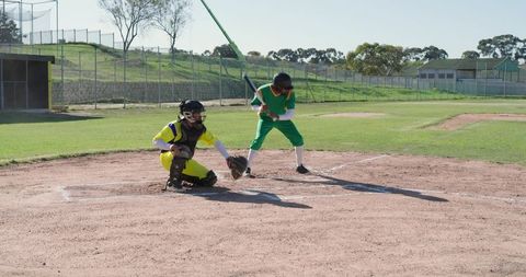 Youth baseball batter swinging at home plate while catcher crouching and reaching on sunny field