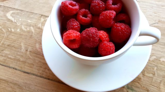 High-angle view of fresh ripe raspberries in a white cup on a wooden table. Perfect for use in food blogs, healthy lifestyle articles, or nutrition-focused advertisements. The vibrant red berries against the neutral tones create an appealing and appetizing image suited for recipes, culinary magazines, or farm-to-table promotions.