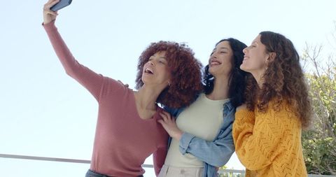 Multicultural women taking sunny balcony selfie in casual denim and knit fall fashion