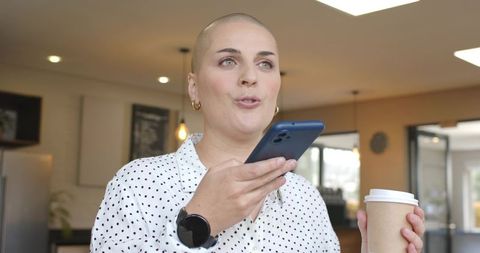 Adult woman speaking into smartphone while holding coffee cup in modern cafe interior