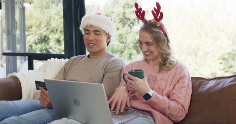 Couple shopping online wearing santa hat and reindeer antlers on cozy sunlit sofa