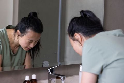 Asian woman leaning over bathroom vanity looking into mirror for morning skincare routine