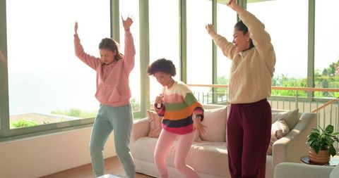 Sunlit diverse friends dancing in seaside living room with floor-to-ceiling windows