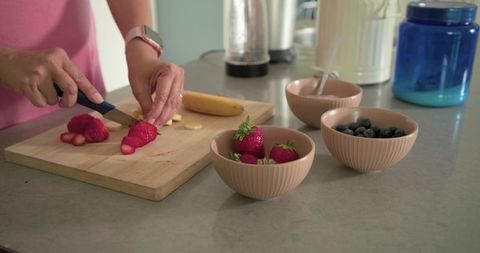 Mature woman slicing strawberries on board preparing healthy breakfast with bowls in pastel kitchen
