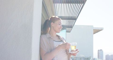 Leaning woman enjoying tea on sunlit high-rise balcony with modern skyline