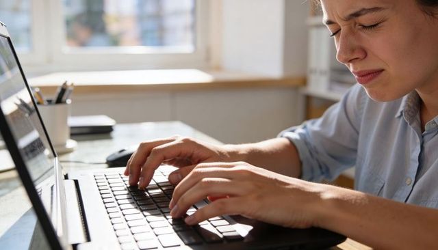 Young woman typing on laptop at sunlit home workspace, squinting from screen glare