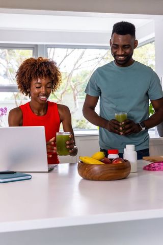 Black couple sipping green smoothies at sunlit modern kitchen island while using laptop