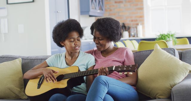 Happy african american mother and daughter sitting on sofa playing guitar. domestic life and quality family time together at home.