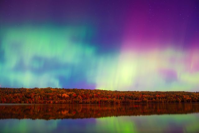 Enchanting display of the Northern Lights over a serene lake reflecting vibrant colors. Forest border in the distance adds a natural touch, perfect for use in travel brochures, nature calendars, desktop backgrounds, or promotional material for outdoor adventures.