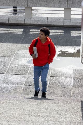 Teen student walking up campus concrete steps in red hoodie carrying notebooks and backpack