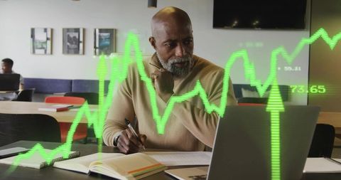 Senior executive reviewing stock charts on laptop and taking notes in modern coworking space