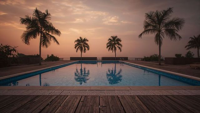 Sunset resort pool reflecting palm trees on wooden deck at oceanfront terrace, serene evening