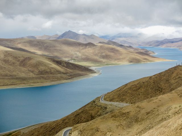 This image captures a winding road along a serene mountain lake bordered by autumn-colored hills. It offers a picturesque and tranquil setting perfect for travel blogs, landscape photography collections, and nature-themed publications. The calm waters and winding road provide a sense of adventure and exploration.