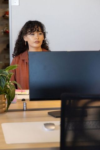 Young professional woman working at modern minimalist office desk behind monitor with plant