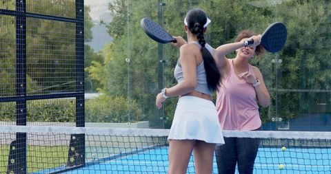 Two african american women playing padel at net on blue court wearing visors outdoor doubles