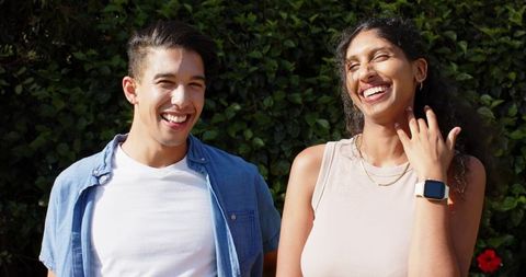 Joyful african american couple laughing in sunlit garden wearing smartwatch and gold chain