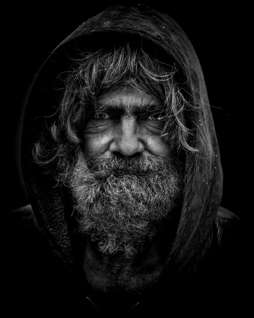 This striking black and white portrait of an elderly man with a bushy beard emphasizes dramatic expressions and deep wrinkles capturing life experiences. The hood adds a mysterious and contemplative essence. Ideal for campaigns on aging, homelessness awareness, or photography exhibitions focusing on human expressions.