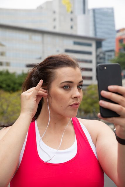 Curvy woman in sports clothes exercising in an urban environment, using her smartphone and putting on earphones. Modern buildings in the background suggest a city setting. Ideal for use in fitness, health, and urban lifestyle promotions.