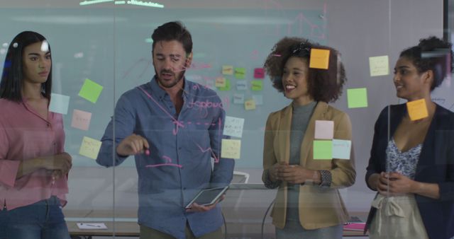 Group of diverse professionals collaborating in an office setting with sticky notes on a glass wall. Ideal for visuals depicting teamwork, strategic planning sessions, creativity in corporate environments, or diverse work teams discussing business strategies.