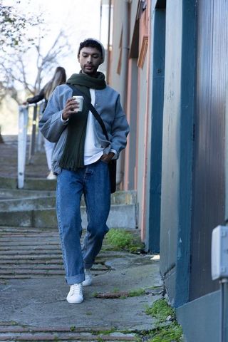 Asian man walking with coffee and crossbody bag on urban stairway wearing beanie and scarf