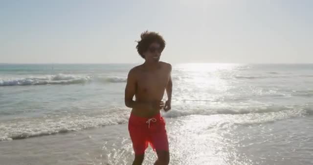 A young, athletic man is running along a sunny beach with waves crashing behind him, highlighting an active and free lifestyle. This setting is suitable for promoting fitness, health, and vacation experiences. Ideal for advertisements related to athletic wear, outdoor activities, or travel destinations.