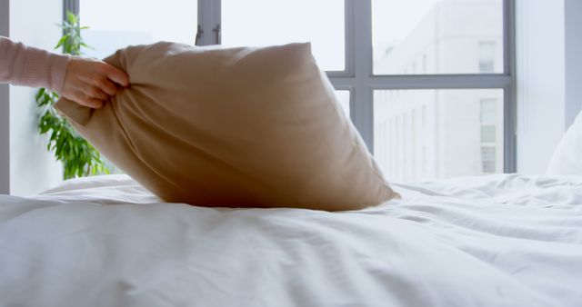 Hand fluffing pillow with beige pillowcase on a bed in a bright modern bedroom with large windows. Ideal for home decor blogs, lifestyle content, and articles about simple living and morning routines.
