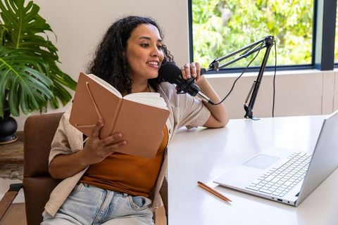 African american woman podcasting at home desk with notebook, laptop, microphone, natural light