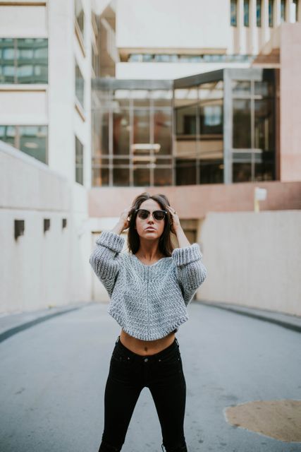 Young woman stands confidently in an urban alleyway, wearing a grey knit sweater, black jeans, and sunglasses. Ideal for use in fashion editorials, lifestyle blogs, or urban style campaigns.