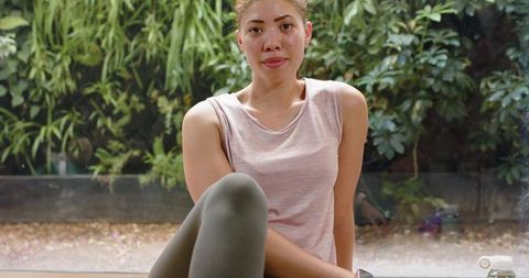 Young woman resting after workout by window with greenery, holding water bottle