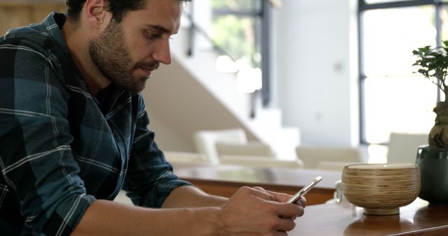 Bearded man in casual plaid shirt using a smartphone while sitting at a wooden table in a modern home interior. Perfect representation of technology usage, casual communication, and contemporary lifestyle themes. Ideal for websites, blogs, or advertisements related to mobile technology, communication, or modern living.