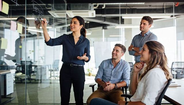 Woman presenting strategy on glass wall while team listening and brainstorming in office