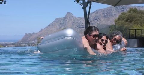 Three women lounging on silver inflatable in infinity pool with mountain and ocean view
