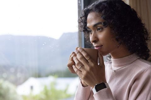 Indian nonbinary person holding mug and gazing out rainy window, cozy introspective moment