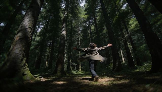 Spinning Man in Olive Coat Dancing Through Sunlit Conifer Clearing with Mossy Trunks