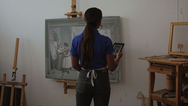 Artist studying framed painting using tablet in studio, wearing cobalt shirt and apron