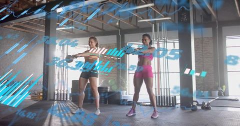 Two women performing kettlebell front raises in industrial gym with blue data overlay