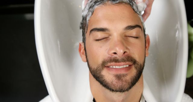 Man with beard relaxing while getting his hair washed at a salon, enjoying the pampered experience. Suitable for promoting salons, spa services, and self-care routines.