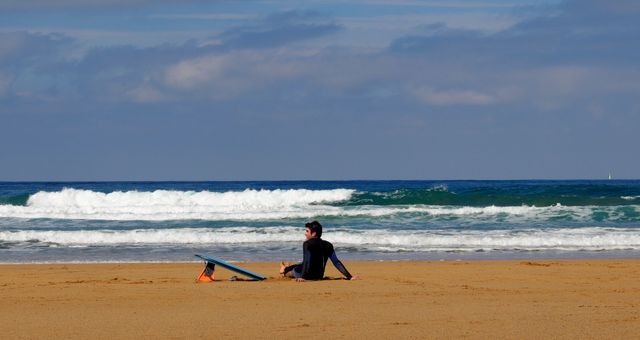This image shows a surfer sitting on the beach with a surfboard while looking at the ocean waves. Ideal for use in content related to surfing, beach holidays, seaside relaxation, water sports, and travel. Suitable for brochures, websites, blogs, advertisements, and social media posts promoting coastal tourism and surfing culture.