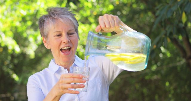 Senior woman wearing casual white shirt pouring refreshing lemon water into glass outdoors. She is smiling and enjoying the drink, surrounded by lush green garden. Ideal for use in advertising about healthy living, hydration, outdoor activities, and wellness.