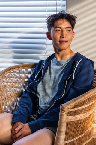 Young asian man sitting in rattan chair bathed in striped sunlight by window blinds